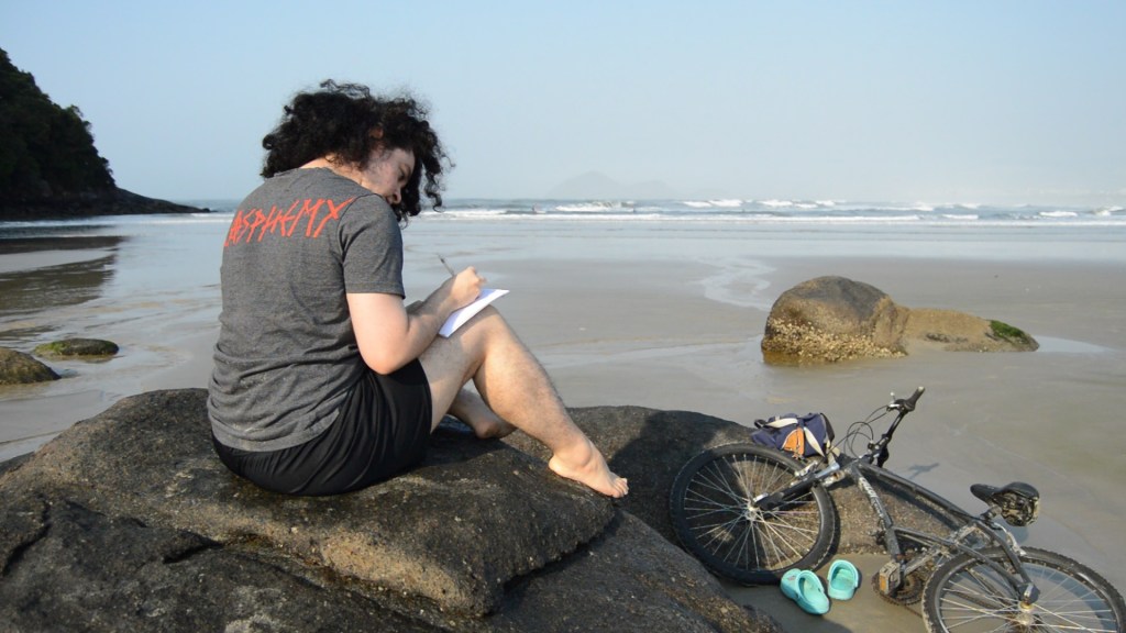 Gionni Blas writing on a piece of paper, sitting on a rock on a beach.