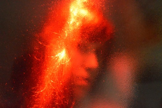 Marcela's face and hair lit by red lighting, behind a glass wall.
