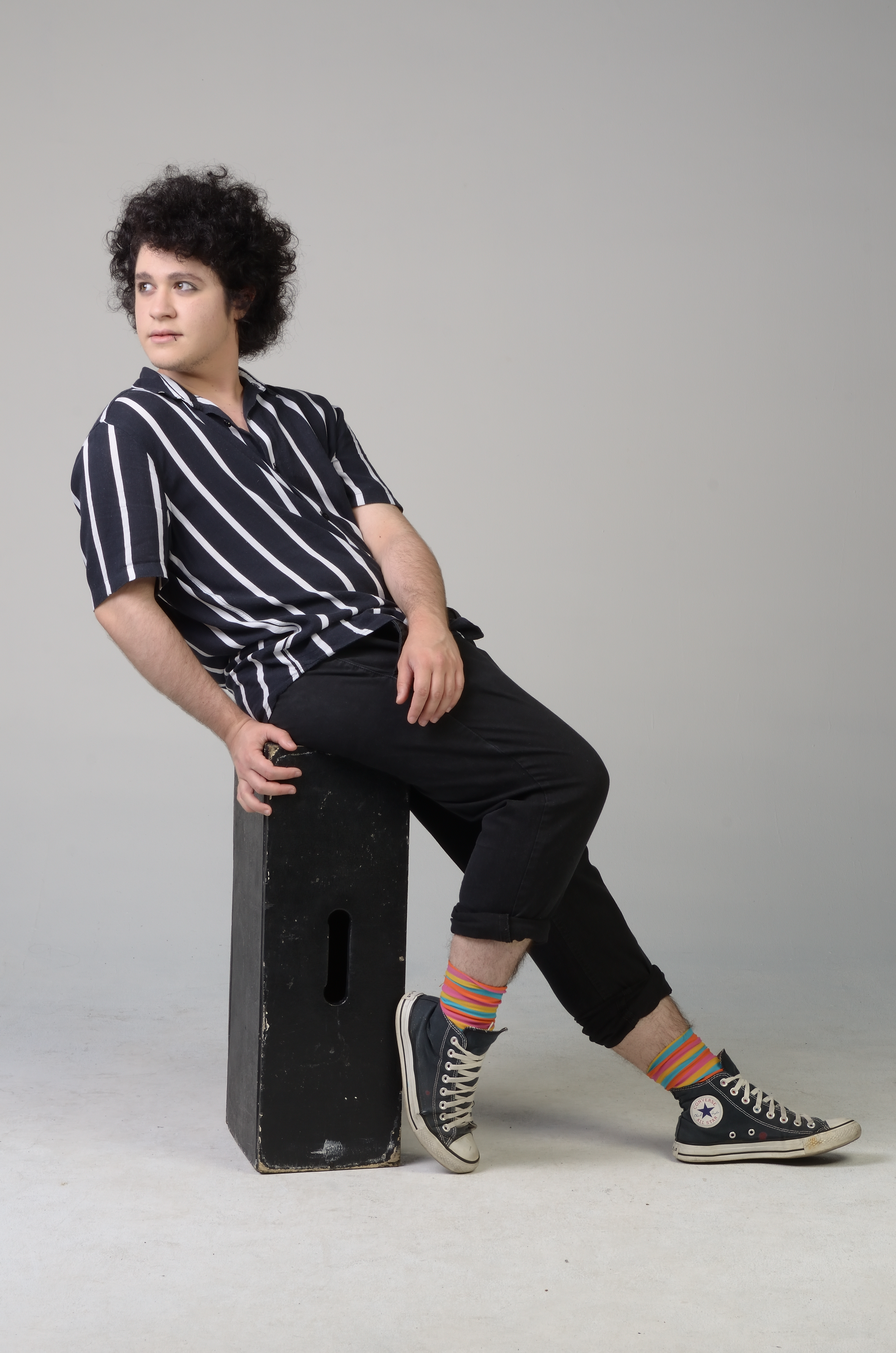 Studio picture of Gionni Blas's full body sitting on a black wooden box in front of a light grey background, leaning and looking to his right. Photographed by Estúdio Thiago Drummond.
