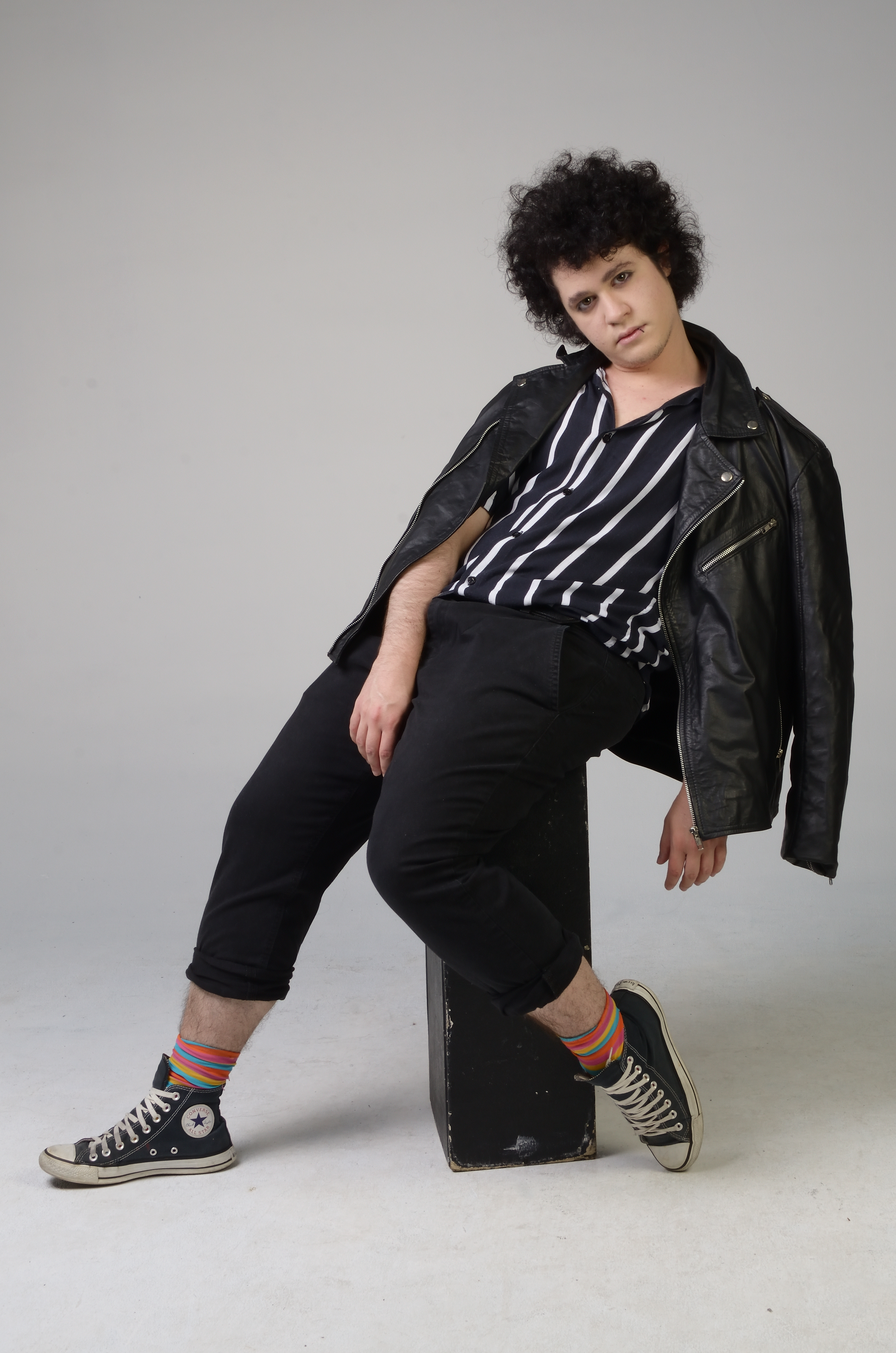 Studio picture of Gionni Blas, full body, sitting on a black wooden box in front of a light grey background, leaning back and looking ahead. Photographed by Estúdio Thiago Drummond.